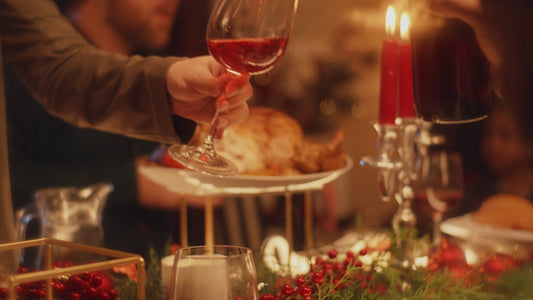 A dining table festooned with greenery and red berries and glowing with warm candlelight. A hand extends a glass of red wine in a toast.